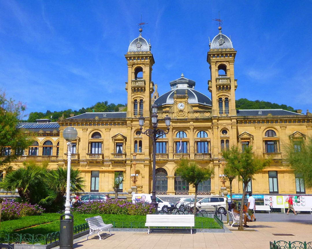 A grand yellow stone building with two towers and a central dome, set in an outdoor plaza with benches, trees, parked cars, and a bright blue sky.